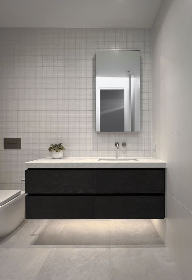 A compact contemporary bathroom featuring a wall-mounted black timber-grain vanity with a white stone benchtop and integrated basin. The back wall is clad in small square light-grey tiles, while the side walls and floor use large pale grey tiles. A rectangular mirrored cabinet is mounted above the basin, with recessed lighting beneath the floating vanity. A partial bathtub and wall-mounted flush plate are visible on the left.