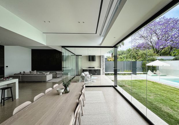 A refined open-plan residential interior with a long dining table in the foreground, a contemporary kitchen and living room beyond, and expansive floor-to-ceiling glazing opening to a landscaped backyard with a swimming pool. The space features a minimalist white ceiling with recessed lighting, dark wall elements, pale timber furnishings, polished concrete-like flooring, and slim black window frames.