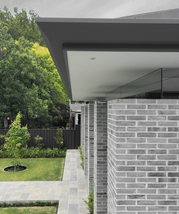 Exterior architectural view of a contemporary single-storey building, focused on a deep roof overhang, crisp white soffit, dark fascia, grey brick walls, and reflective glazing. A paved walkway, lawn, planted garden, and dark boundary fence frame the building.