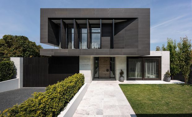 Modern two-storey residential facade with a dramatic projecting upper-level box clad in dark grey panels, deep recessed glazing with vertical fins, and a lighter ground floor. A paved path leads to the entry, with a dark gate, manicured hedge, lawn, and potted plants.