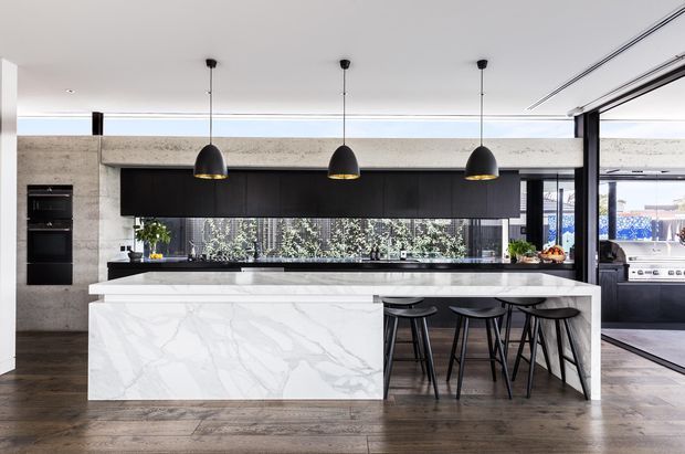 Contemporary open-plan kitchen with a large white marble-look island and integrated seating for four. The space features dark timber or black cabinetry, a long horizontal splashback window, exposed concrete-like wall surfaces, pendant lighting, dark timber flooring, and direct access to an outdoor barbecue area through large glazed openings.