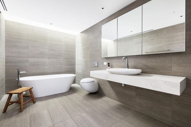 Contemporary minimalist bathroom with large-format taupe-grey tiles covering the walls and floor. The room features a freestanding white bathtub, wall-mounted white vanity with integrated basin, floating mirrored cabinetry, a wall-hung toilet, and restrained chrome fixtures. A small timber stool adds warmth to the otherwise neutral space.