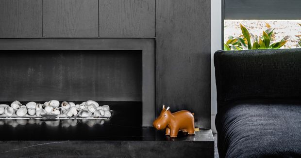Close-up of a dark contemporary living room fireplace with a charcoal textured surround and black hearth. White decorative stones sit within the fireplace, while a small orange animal figurine rests on the hearth beside a dark upholstered sofa. A narrow window or opening behind the sofa brings in daylight and greenery.
