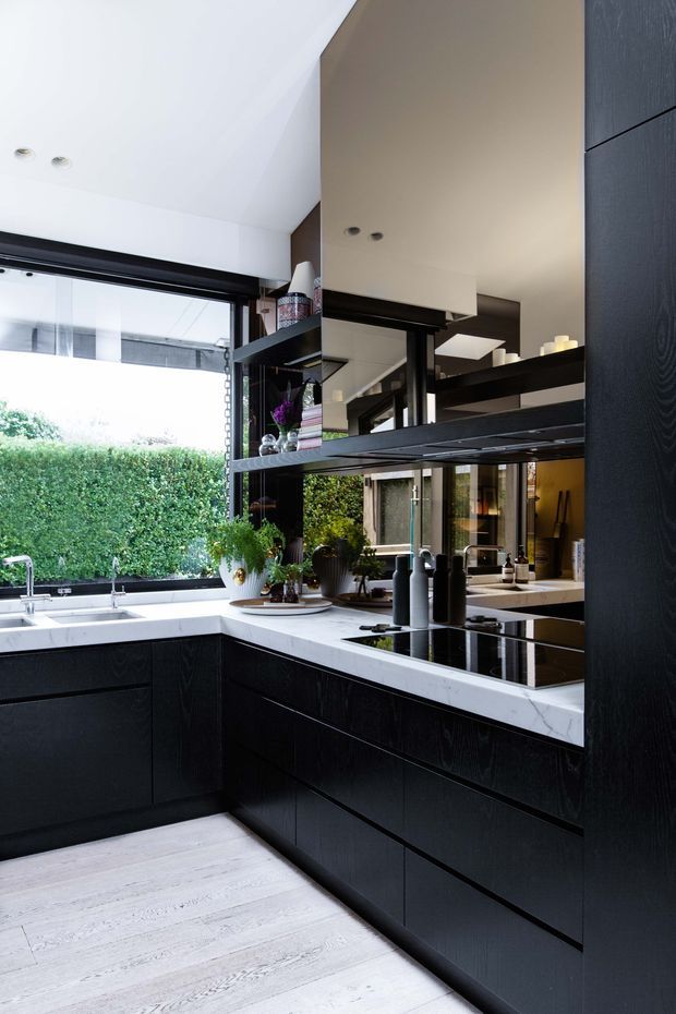 A contemporary L-shaped kitchen with matte black wood-grain cabinetry, a white marble-look benchtop, integrated black induction cooktop, stainless-steel sink and tapware, and open black shelving. Large glazing opens the space toward a lush green hedge, while mirrors and reflective surfaces visually extend the room. The pale timber-look floor and white ceiling create a bright contrast with the dark joinery.
