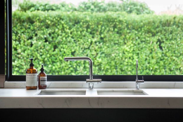 Close-up of a contemporary kitchen sink area with a white marble-look benchtop, double undermount sink, chrome mixer tap and separate filtered-water tap. A large black-framed window overlooks a dense green hedge, with amber soap dispensers positioned on the counter.