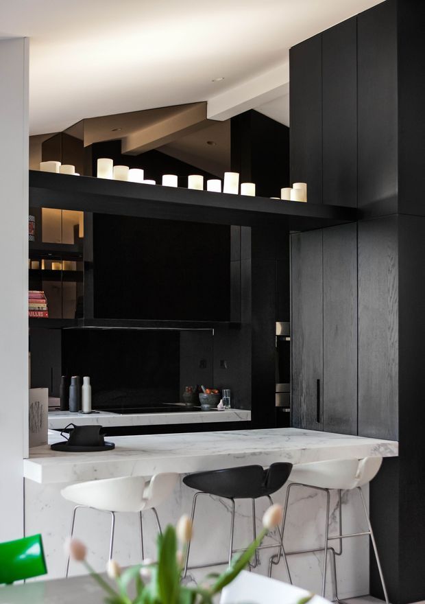 A dramatic, contemporary kitchen with extensive matte black cabinetry, a black tiled or stone backsplash, and a large white marble island with integrated seating. The island has a waterfall edge and is paired with white and black bar stools. Open shelving, pendant-style candle-like lights, dark reflective surfaces, and exposed structural ceiling beams create a refined, moody atmosphere.
