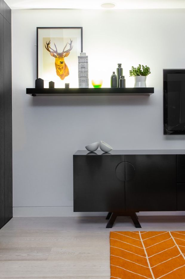 A styled contemporary living room vignette featuring a black low-profile media console, floating black display shelf, framed deer artwork, decorative vessels, a potted plant, and a wall-mounted television. Pale timber flooring and a vivid orange geometric rug contrast with the predominantly white and charcoal palette.