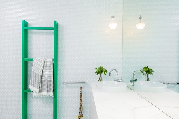 Bright contemporary bathroom with a white marble-look double vanity, vessel basins, chrome faucets, large mirrored wall, globe pendant lights, and a vivid green ladder-style towel rack holding striped towels. A subtle textured wall surface and small potted plants add detail.