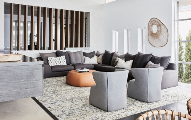 Bright, contemporary living room with a large charcoal-grey sectional sofa, two light-grey lounge chairs, and a rounded tan leather ottoman arranged on a textured area rug. White walls, tall windows, and a vertical timber-and-black divider add architectural interest, while woven wall art and neutral furnishings create a refined, airy atmosphere.