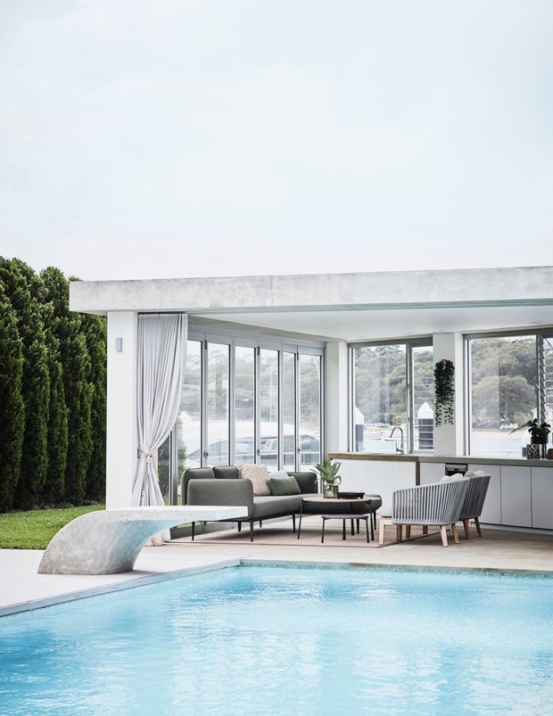 A bright modern poolside living area with a broad concrete roof slab, extensive floor-to-ceiling glazing, white curtains, and a minimalist indoor-outdoor lounge. A swimming pool dominates the foreground, while a grey sofa, coffee table, armchairs, and a sleek kitchen or bar counter sit beneath the covered terrace. The setting is landscaped with clipped greenery and views beyond the glazing.