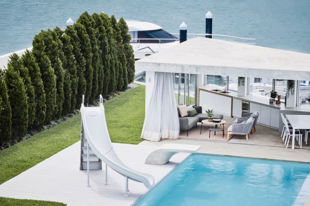 A bright, modern waterfront residence with a flat-roofed pavilion opening onto a swimming pool and pool deck. The outdoor living area includes a sofa, armchairs, coffee table, curtains, and an adjoining dining and kitchen space. A large waterslide leads into the pool, while sculpted evergreen hedging provides privacy along the waterfront edge.