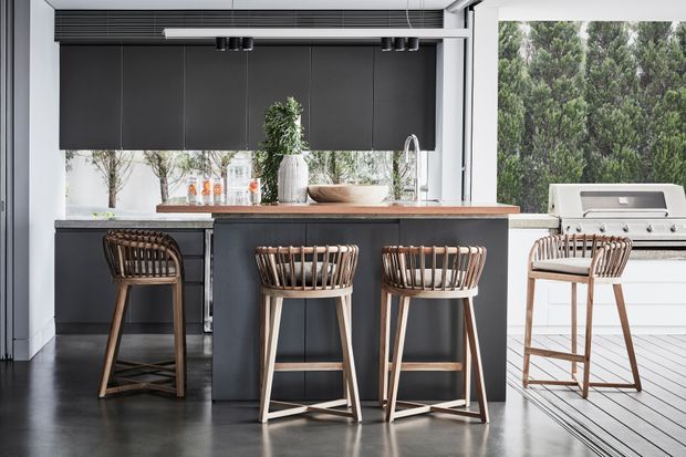 A contemporary open-plan kitchen with a dark island, integrated cabinetry, timber bar stools, stainless-steel appliances, and a timber benchtop. Large glazed openings bring in natural light, with an outdoor barbecue visible to the right.