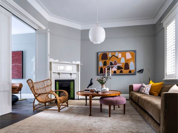 A refined living room with grey walls, ornate white ceiling cornices, a traditional fireplace, plantation shutters, and an open connection to an adjacent room. The space combines a rattan lounge chair, brown sofa, round timber coffee table, upholstered ottoman, contemporary pendant light, and colourful abstract artwork over a large neutral rug.
