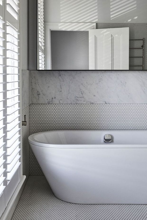Bright, compact bathroom featuring a freestanding white bathtub, small-format circular mosaic tiles across the floor and lower walls, a pale veined stone feature band, white painted trim and door, and plantation shutters. A large mirror reflects the doorway and tiled wall, while strong daylight creates horizontal shadow patterns.