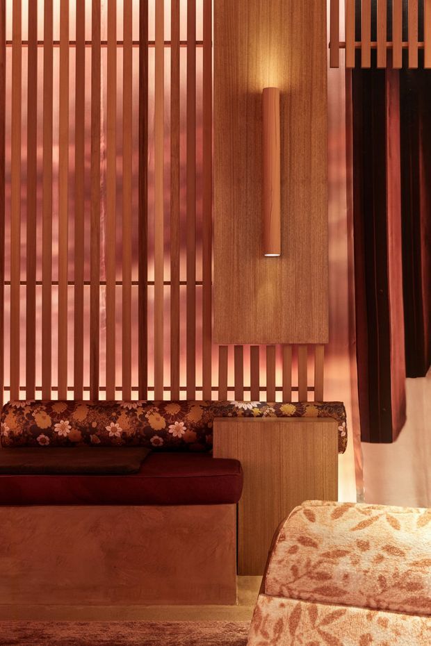 A close-up interior view of a richly layered seating area, featuring warm timber vertical slats, a timber wall panel with an integrated elongated wall light, built-in cabinetry, and upholstered seating. The palette is dominated by reddish brown timber, burgundy upholstery, patterned floral cushions, and warm ambient lighting. The composition has a retro, richly textured character with strong linear screening and custom joinery.
