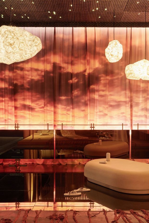 A dramatic, atmospheric interior lounge or hospitality space with a reflective dark floor, warm amber-red translucent wall panels, and a richly patterned perforated ceiling with recessed spotlights. Large sculptural cream pendant lights hang overhead. Rounded cream upholstered seating and low tables sit in front of mirrored or highly reflective surfaces, creating layered reflections. The space has a luxurious, moody, theatrical character with strong warm lighting.