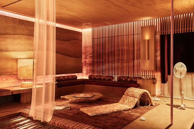 A warmly lit, richly timber-lined lounge or relaxation room with low built-in seating arranged around a dark rug and low coffee table. Vertical timber screening divides the space, while sheer curtains, sculptural lounge furniture, textured cushions, and an integrated wall light create an intimate, luxurious atmosphere. The room has a low, standard-height ceiling and a strong Japanese-inspired spa or retreat character.