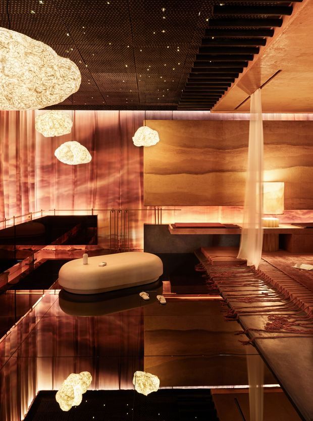 A dramatic, luxurious lounge or living room with a warm amber palette. The space features reflective dark flooring, a sculptural cream upholstered coffee table or ottoman, low dark seating, layered rugs, translucent drapery, large textured wall panels, and sculptural pendant lights. A dark perforated ceiling with numerous pin lights and a striking black linear ceiling feature create a theatrical atmosphere. The image appears vertically mirrored or reflected, extending the room visually.