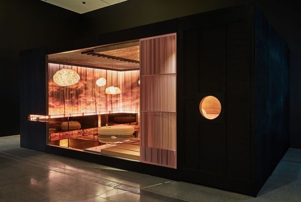 A dark, dramatic interior installation resembling a luxury lounge or hospitality space, viewed from outside through a large glazed opening. The space features warm amber lighting, reflective dark flooring, richly patterned illuminated wall surfaces, pendant lights, built-in seating, and a striking vertical slatted screen. A circular illuminated opening is visible in the dark surrounding wall.