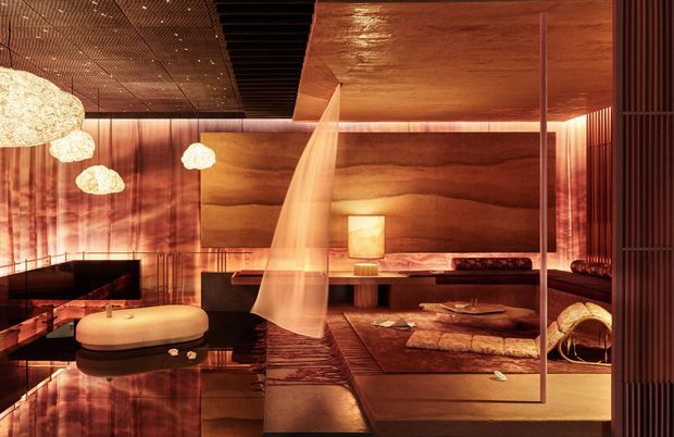 A richly layered, dimly lit contemporary living room or media room with extensive warm timber wall and ceiling surfaces, dark reflective flooring, a large low black sofa, sculptural lounge seating, a television, textured rugs, and a suspended translucent curtain or screen. Organic pendant lights and red-orange ambient lighting create an intimate, luxurious atmosphere. Vertical timber battens and a large framed timber feature wall add depth and architectural detail.