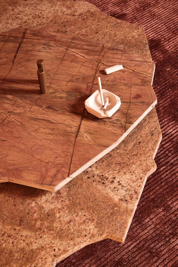Close-up product-style photograph of layered, irregularly shaped stone or stone-look coffee tables with richly veined brown tops and speckled tan surfaces. The tables sit on a dark rust-brown textured carpet and are styled with a small ashtray, cigarette, lighter, and cylindrical object. Warm, moody vintage-inspired composition.