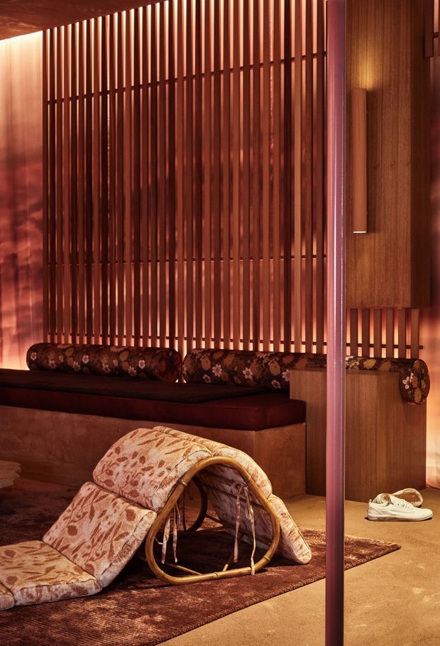 Warmly lit interior lounge featuring a low built-in upholstered bench with floral cylindrical cushions, a sculptural curved lounge chair draped in patterned fabric, and a richly textured rug. The dominant architectural feature is a full-height timber slatted screen with integrated warm lighting, complemented by dark timber wall panelling and a slim vertical structural or decorative post. The space has a moody, intimate, retro-inspired atmosphere.