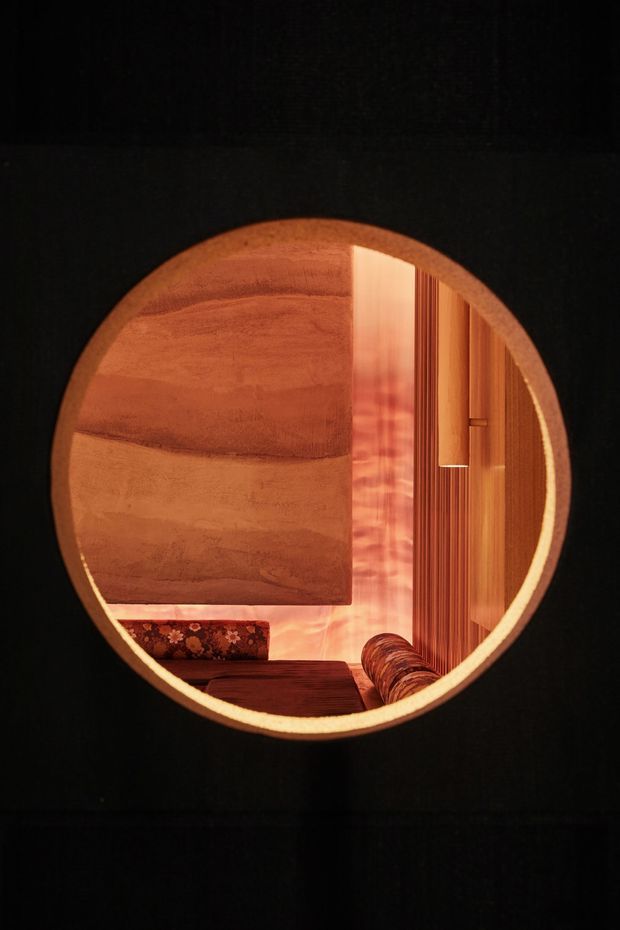 A dark, atmospheric interior view framed through a large circular opening. Inside is a warm wood-lined lounge or relaxation space with vertical timber slats, a curved upholstered seating area, a patterned cushion, and a softly glowing amber-lit wall or ceiling surface. The composition emphasizes circular geometry, deep shadows, and rich reddish-brown timber tones.