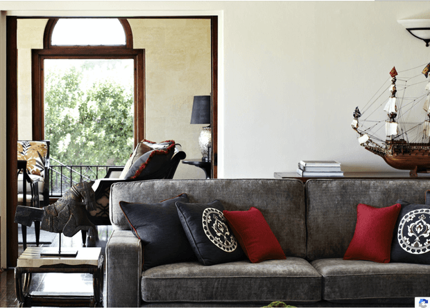 A refined living room centered on a large charcoal-grey sectional sofa with black and deep red cushions. Dark timber-framed glazing and an arched transom window open to a leafy balcony or exterior view. A black lounge chair, side table, lamp, books, and a decorative model ship complete the room. The space combines neutral finishes with rich red and dark timber accents.