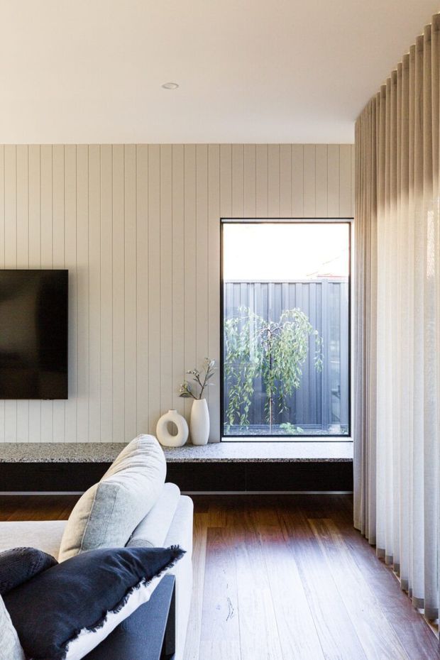 Bright contemporary living room with a wall-mounted television, built-in low black cabinetry, pale vertical wall panelling, timber flooring, a light upholstered sofa, and full-height sheer curtains beside a large window. Neutral finishes and warm natural light create a clean, refined atmosphere.