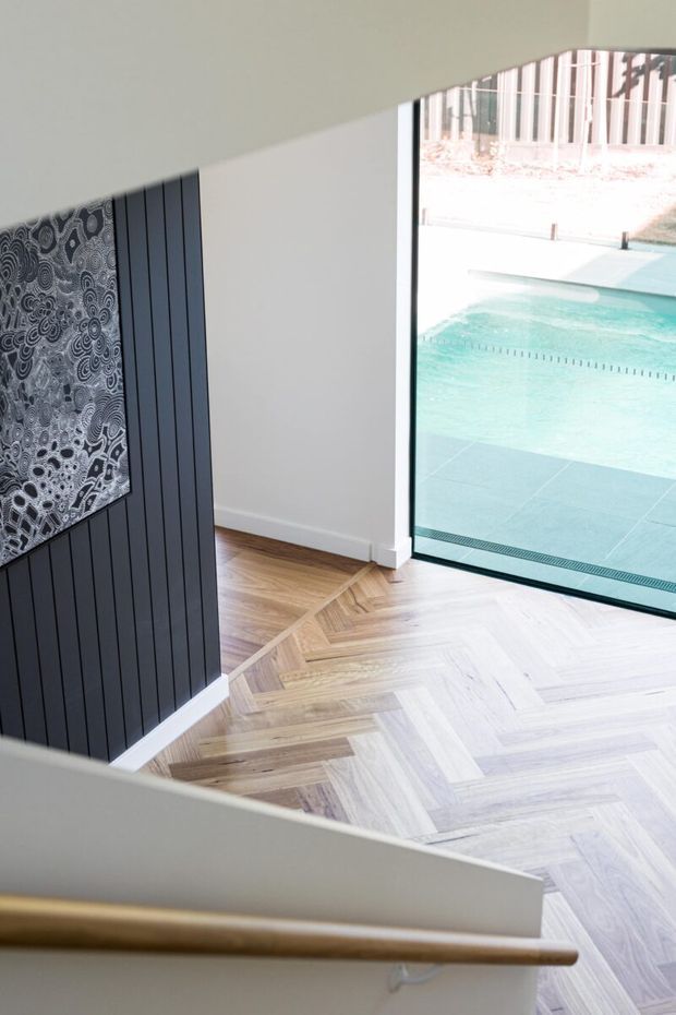 Interior view from a stairway or landing, showing a light timber herringbone floor, dark vertical wall panelling, a timber handrail, and large black-framed glazing opening toward a swimming pool. A patterned artwork is mounted on the feature wall.