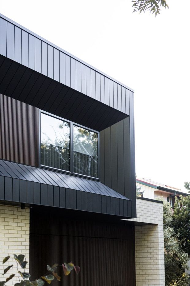 A close-up exterior architectural photograph of a contemporary multi-level home. The facade combines dark vertically seamed metal cladding, a projecting black metal canopy and roof edge, vertical timber-look screening, large dark-framed windows, and pale cream brickwork at the lower level. Mature foliage partially frames the building, while a neighbouring roof is visible in the background.