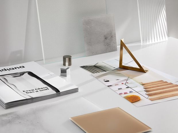 A bright, minimalist materials and design sample arrangement on a white worktable, featuring architectural magazines, tile and surface samples, printed material boards, a small metal cylinder, and a geometric wooden display stand. White textured panels and vertical ribbed detailing form the background.