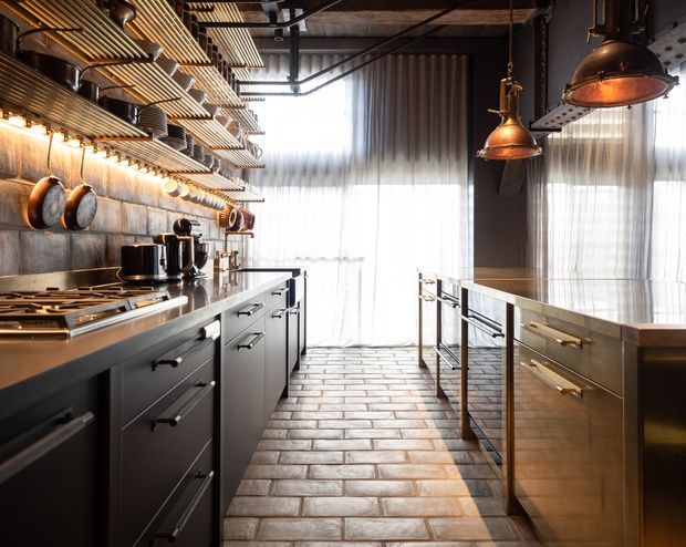 A narrow, industrial-style kitchen with dark built-in cabinetry, stainless-steel benchtops and appliances, exposed ceiling services, metal shelving, hanging copper pendant lights, and cookware displayed along a tiled wall. A central island and long galley-style layout lead toward full-height sheer curtains and bright glazing at the rear. The floor is finished in warm rectangular brick-like tiles, creating a rugged commercial-kitchen atmosphere.