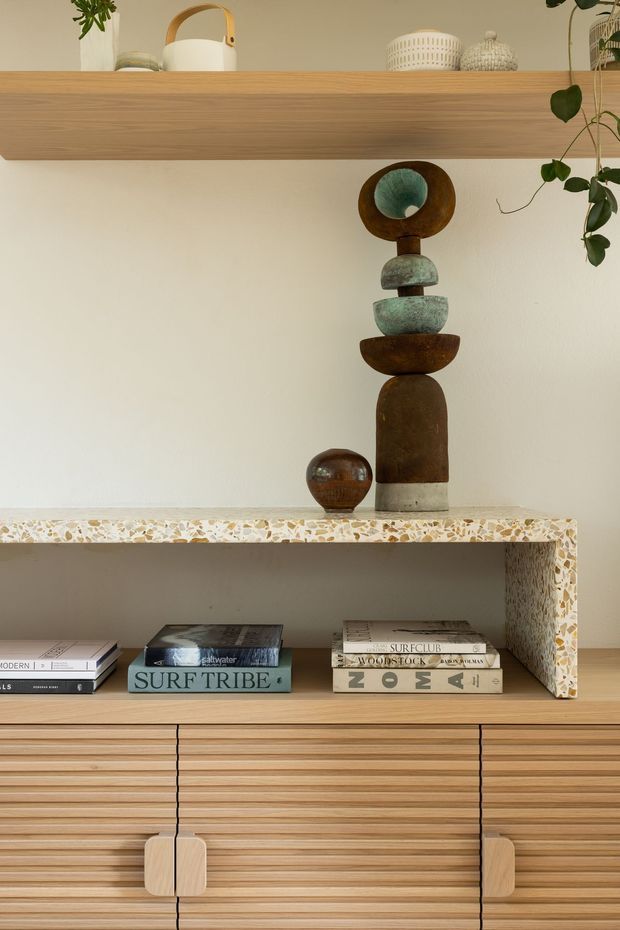 Close-up interior detail of a warm timber built-in shelving and cabinetry unit. A long floating timber shelf displays ceramics, plants and sculptural objects above a terrazzo-edged open niche with books, while the lower section features vertically grooved timber cabinet fronts with rounded timber handles. The palette is warm and neutral with natural decorative accents.