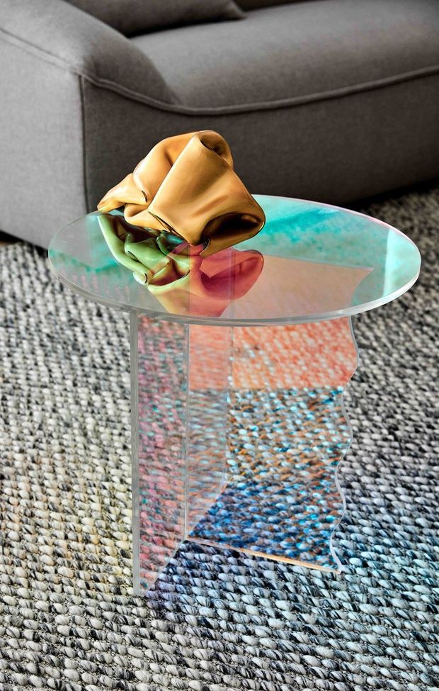 Close-up interior photograph of a sculptural transparent glass side table with a round top and curved pedestal base, positioned on a textured grey woven rug beside a charcoal-grey upholstered sofa. A colourful abstract decorative object sits on the tabletop, with strong reflections visible through the glass.