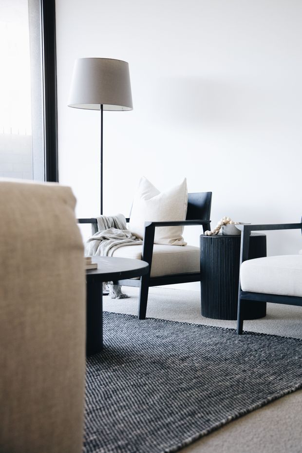 A bright, minimalist living room vignette with black-framed lounge chairs, a neutral upholstered sofa or armchair, white cushions, a slim floor lamp, a round dark side table, and a textured charcoal rug over pale carpet. Large black-framed glazing and white walls create an airy, refined atmosphere.