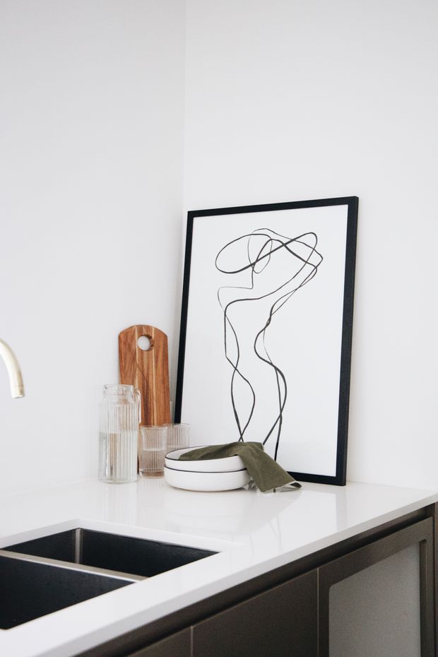 A close-up of a minimalist kitchen corner featuring a white countertop with an inset dark double-basin sink, dark cabinetry, and a simple black-framed abstract line artwork leaning against the wall. Clear ribbed glass vessels, a wooden serving board, and a shallow ceramic bowl with an olive-green cloth add restrained decorative detail.