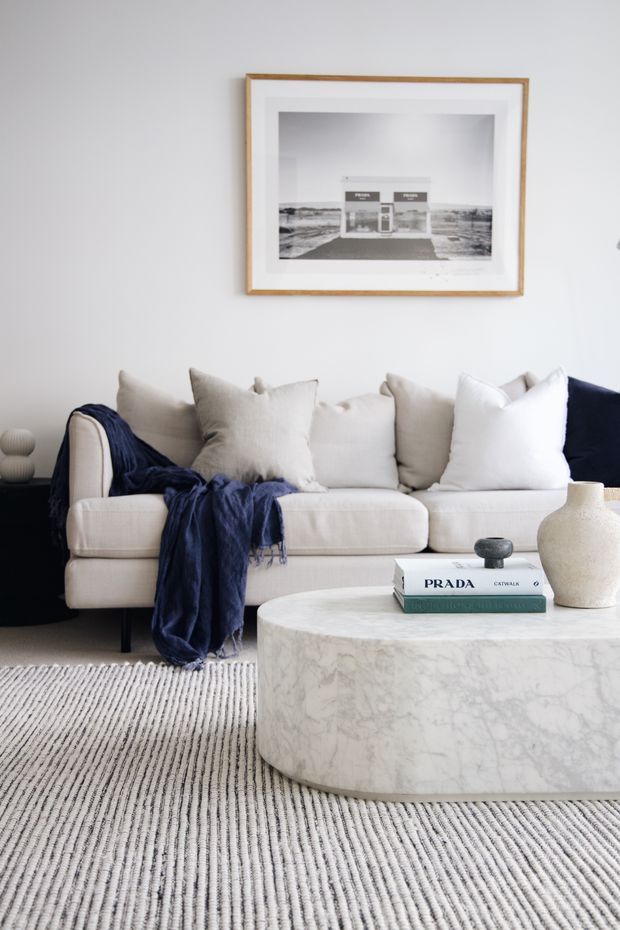 A bright, refined living room featuring a large cream upholstered sofa layered with neutral cushions and a deep navy throw. A rounded white marble coffee table sits on a textured striped rug, with a ceramic vase and stacked books. The white wall has a simple light timber-framed monochrome artwork.