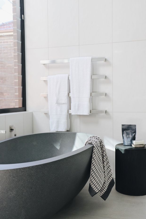 Bright contemporary bathroom featuring a large freestanding dark stone bathtub, white tiled walls, a black-framed window, wall-mounted towel rails with white towels, and a dark ribbed cylindrical side table or cabinet. The space has a clean, minimalist monochromatic palette with natural daylight.