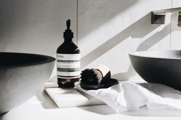 Close-up of a minimalist bathroom vanity styled with two dark stone vessel basins, a wall-mounted chrome tap, a pale tiled or plastered wall, and a white countertop. An Aesop hand wash bottle, soap dish, and folded white towel add functional details. Strong directional sunlight creates crisp shadows across the clean, monochromatic space.