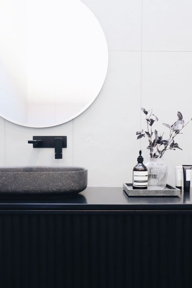 Close-up of a refined contemporary bathroom vanity. A large round frameless mirror hangs above a dark stone vessel basin, with a matte black wall-mounted tap and black fluted vanity cabinetry below. Pale large-format wall tiles provide a clean backdrop, while a tray with monochrome toiletries and a glass vase adds styling.