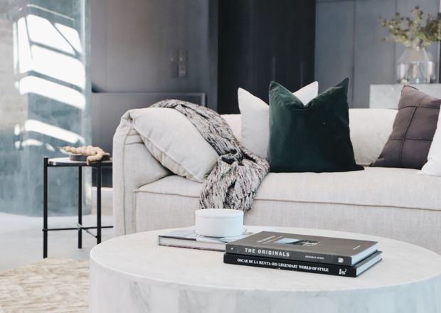 A close-up view of a softly styled contemporary living room, featuring a pale upholstered sofa with layered white and charcoal cushions, a dark green accent cushion, and a textured throw. A round light stone or marble coffee table holds stacked books and a cup, while a small black side table and large window add depth. The palette is cool and neutral with refined, cozy styling.