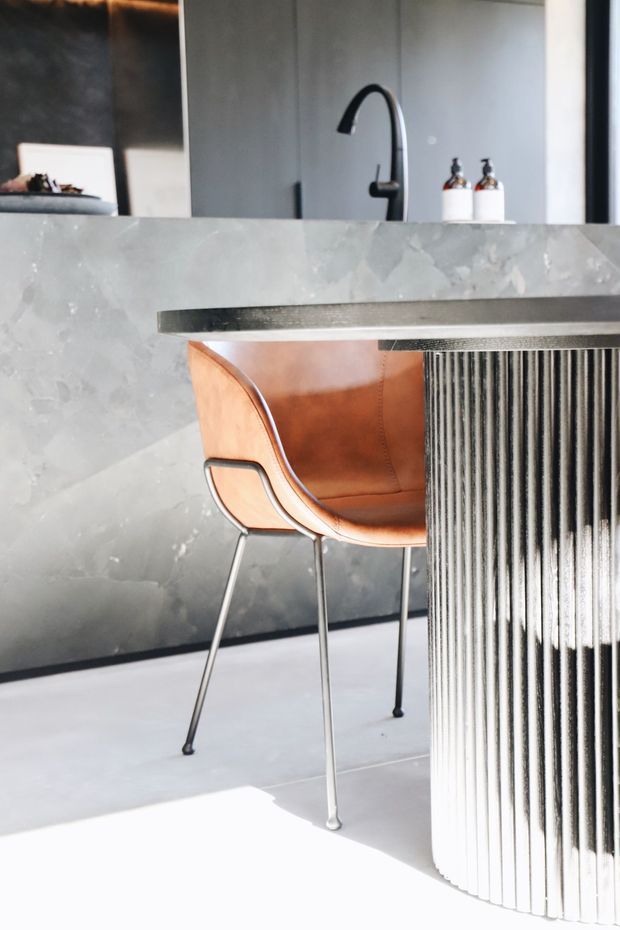 Close-up interior view of a contemporary kitchen or entertaining area, featuring a grey stone island with a fluted cylindrical support, a dark benchtop, black gooseneck tap, integrated sink area, and a tan leather bar chair with slim metal legs. The palette is monochromatic grey and white with warm leather contrast and strong sunlight.
