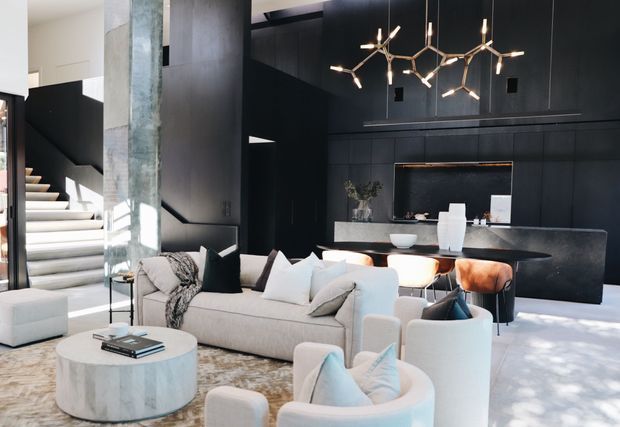 A sophisticated open-plan living room with a dramatic dark feature wall, integrated bar or kitchen joinery, sculptural pendant lighting, a pale sectional sofa, curved lounge chairs, and a round stone coffee table. A staircase and tall glazed opening are visible to the left. The interior has strong contrast between black built-in surfaces and light upholstery, with abundant natural light.