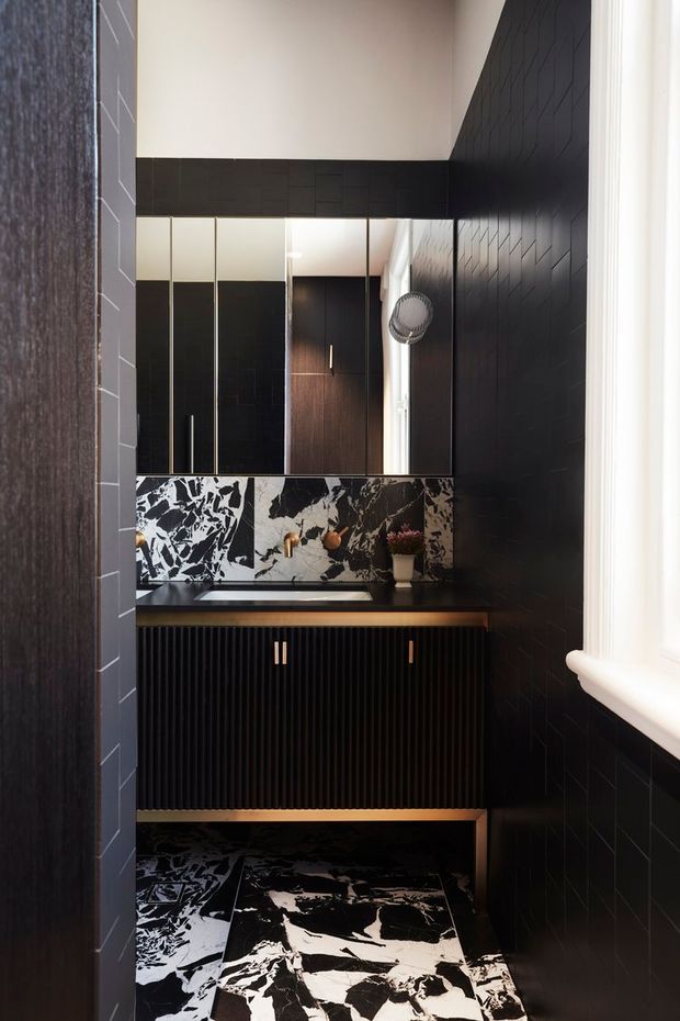 A compact, dramatic bathroom with black wall tiles, mirrored upper cabinetry, a dark vanity with vertical fluted detailing and brass-toned hardware, and bold black-and-white marble-patterned splashback and floor tiles. Natural light enters from the window on the right, contrasting with the moody dark finishes.
