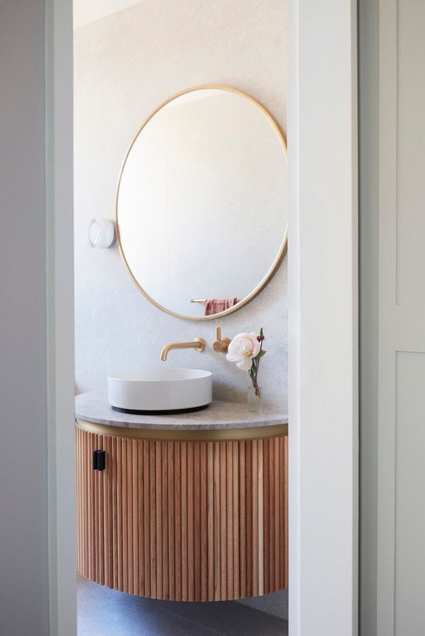 A compact, refined bathroom vanity viewed through a doorway. The space features a round vessel basin on a curved stone benchtop, a fluted timber vanity with a curved front, a large round mirror with a slim brass-toned frame, brushed brass tapware, and pale textured wall finishes. The styling is light, warm, and contemporary.