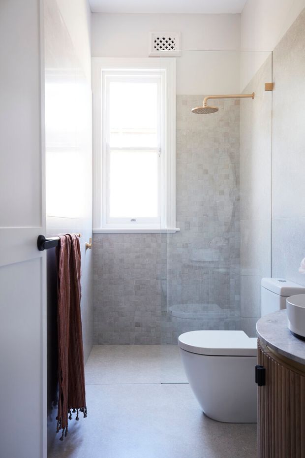 Bright, compact contemporary bathroom viewed through a partially open door. The room features a walk-in shower with a frameless glass screen, pale textured mosaic wall tiles, a white toilet, a small window, a wall vent, and a curved stone vanity with fluted timber cabinetry. A dark towel rail with a hanging textile is visible near the entrance.