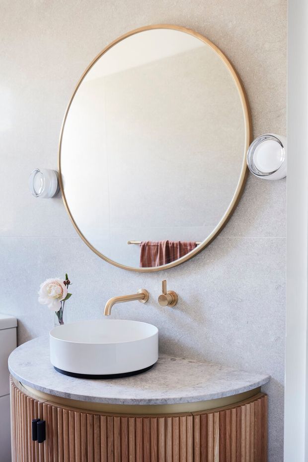 A refined bathroom vanity featuring a round vessel basin on a curved stone countertop, vertically fluted timber cabinetry, a wall-mounted brushed brass tap, and a large round mirror with a timber-toned frame. The pale textured wall and soft neutral palette create a warm, elegant look.