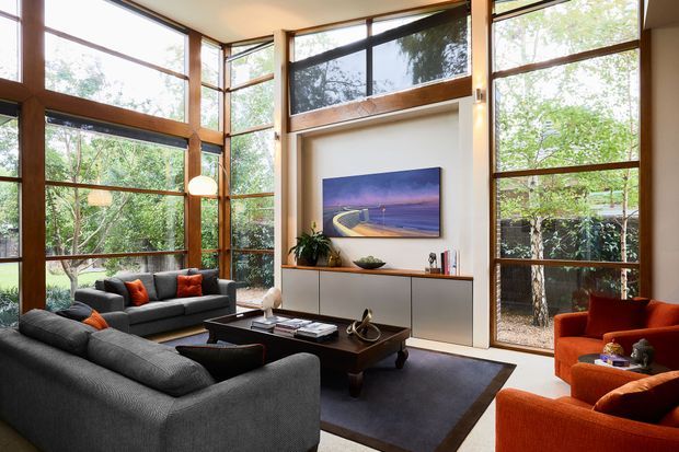 A spacious contemporary living room with floor-to-ceiling timber-framed glazing on multiple sides, overlooking dense greenery. Two grey sofas and vivid orange lounge chairs surround a dark coffee table and rug, while a built-in wall unit, artwork, and indoor plants complete the warm, light-filled interior.