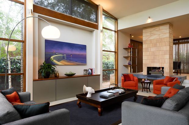 A spacious contemporary living room with floor-to-ceiling timber-framed glazing, a large tiled fireplace feature wall, built-in dark cabinetry, and a low dark wood coffee table. Grey sofas and orange-red lounge chairs are arranged around the fireplace, with a large sculptural pendant light and artwork adding visual interest. The room has a warm, refined mid-century-influenced atmosphere and strong indoor-outdoor connection through the extensive windows.
